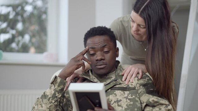 Frustrated military man looking at picture as loving woman talking calming down spouse. Portrait of desperate African American handsome husband and caring Caucasian beautiful wife at home indoors
