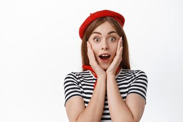 Close up of amazed young woman student in red beret, gasping excited and surprised, holding hands on cheeks, checking out banner, big news, white background