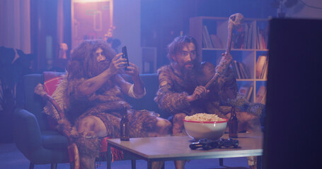 Obraz premium Humor concept. Ancient homo sapiens couple of cavemen in savage animal fur drinking beer eating popcorn watching movie in dark smoke living room.
