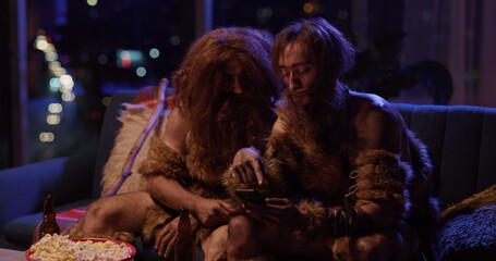 Fototapeta premium Funny neanderthals using mobile phone in modern house living room. Ancient prehistoric couple of two male friends wearing animal fur playing video games.