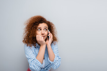 confused woman looking away while talking on smartphone isolated on grey