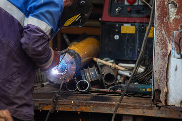 a man in a welder's mask and in a robe works in the factory, is engaged in welding work, the flow of hot plasma welds the seam on a metal pipe, production, plant and rough work

