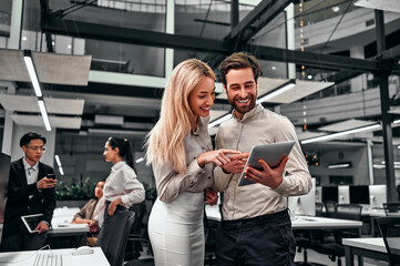 Two beautiful successful smiling business people are standing looking at the tablet screen in a spacious office. Colleagues are working in the background.