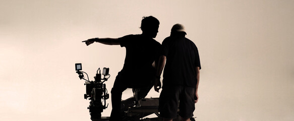 Behind the scenes of film production. Silhouette camera man and film crew setting up studio for recording or shooting. Videographer with 4K UHD camera and tripod stand in studio production. 