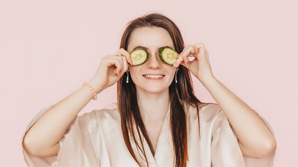Girl make homemade face beauty masks. Cucumbers for the freshness of the skin around the eyes. Woman take care of youthful skin. Model laughing and having fun in spa on pink background