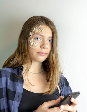 A happy cheerful teenage girl with a picture on her face of flowers holds a smartphone in her hand, looks at the phone screen, uses the Internet