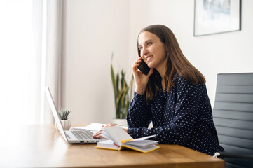 Executive female office employee takes a phone call sitting on the workplace, smiling young woman has pleasant phone conversation, talking and chatting on the smartphone in the office