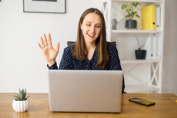 Cheerful businesswoman is holding video call on the laptop computer, sits in the modern office and greeting with customers or colleagues, has video meeting on the distance, involved online conference