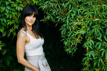 Portrait of a young sexy brunette in a white light T-shirt near green leaves of grapes.