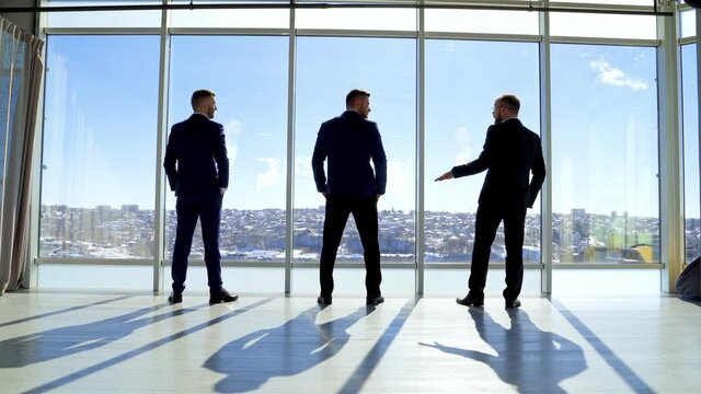 Business people in office on city landscape through window. Three men in elegant suits standing by the window and talking in sunny daytime. Back view.