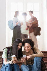 African student with coffee to go sitting near african american friend using smartphone