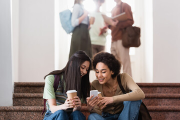 Smiling multiethnic students with coffee to go using cellphone