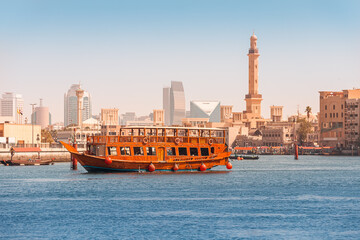 An authentic wooden cruise ship cruises and transports tourists along the Dubai Creek Canal with skyscrapers and minarets of mosques in the background. Travel and sightseeing
