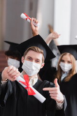 Graduate in medical mask pointing at blurred diploma