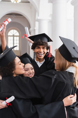 Cheerful multiethnic students with diplomas embracing in university