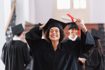 Cheerful african american graduate in academic dress holding cap and diploma