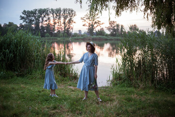 Naklejka na ściany i meble Woman posing outdoor in the nature with bouquet of flowers and the gorgeous view of lake and sun. the mother having fun with girl or daughter. Models dressed in blue dresses. Happy mothers day