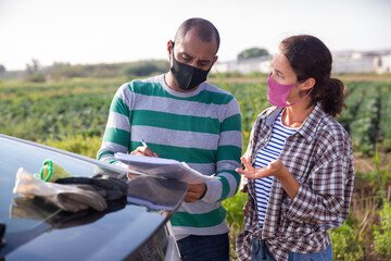 Woman and man farm workers wearing face masks for spread viral disease discussing papers standing near car outdoors