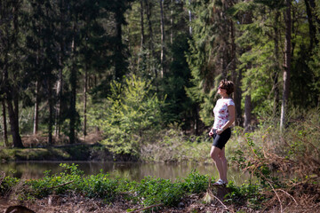 Junge schlanke dunkelhaarige Frau mit kurzen Haaren, steht auf der rechten Seite vom Bild vom See im Wald, schaut in die Ferne und denkt nach. Auf der linken Seite ist viel Platz. Es ist Sommer. 