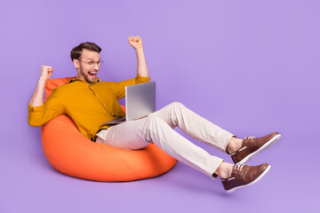 Portrait of attractive cheerful guy sitting on chair using laptop rejoicing isolated over violet purple color background