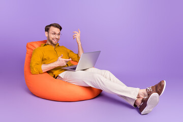 Full length body size photo of guy sitting in beanbag using laptop having video meeting isolated on vivid violet color background