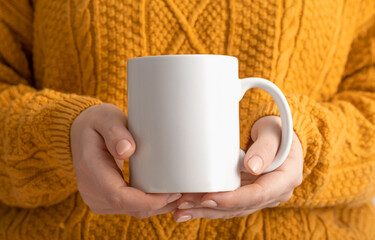 Female hands hold mock up white empty mug, cup for your design and logo close-up. Woman in yellow knitted sweater autumn winter. Blank template for promotional text message or promotional content.