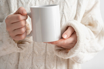 Female hands hold mock up white empty mug, cup for your design and logo close-up. Woman in beige warm knitted sweater autumn winter. Blank template for promotional text message or promotional content.