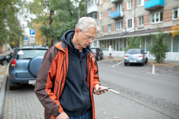 A elderly gray-haired man with a smartphone in a city parking lot. Car parking mobile payment concept