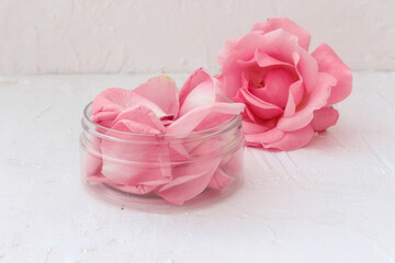 idea for natural rose oil cosmetics. Beauty jar filled with natural rose petals standing together with rose flower on textured white background