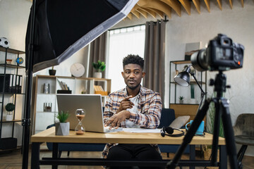 Young afro-american streamer in casual outfit sitting at table and using professional video camera,...