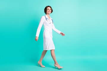 Full length body size side profile photo female doctor walking forward wearing white uniform isolated vivid teal color background
