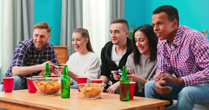 Multiethnic people celebrate video game victory in home living room. Diverse friends enoying beer and chips. Excited friends playing video games at home and having fun