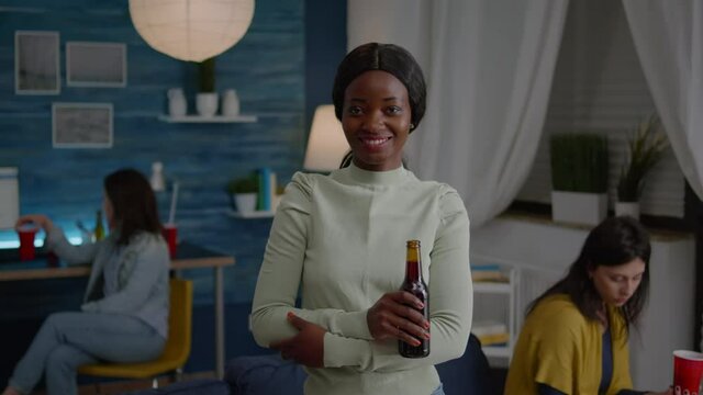 Potrait of afro american woman smiling into camera while holding beer bottle in hands late at night in living room. In background multiethnic friends gathering together having fun during wekeend party