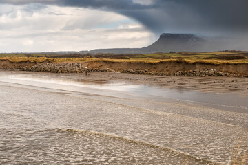 Rosses point beach and Benbulben flat top mountain covered in snow in county Sligo, Ireland, Warm sunny day, cloudy blue sky covers the peak of the mountain.