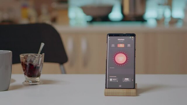 Mobile phone with wireless lighting automation software placed on kitchen desk in empty house with smart system, turning on lights. Smartphone with high tech app controlling electricity effiency