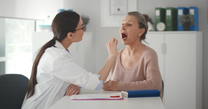 Female doctor checking throat of senior patient while examining her during consultation in clinic