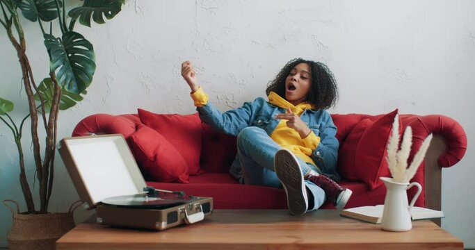 Cool stylish african american black female in yellow hoodie and jeans jacket listening music on vinyl turntable vintage record player dancing sitting on red sofa at home. Cheerful woman. Urban style
