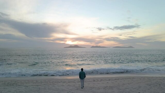 Video zoom in de un hombre caminado hacia el mar en una playa a la puesta de sol con islas al fondo.