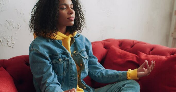 Stylish african american black female in yellow hoodie jeans jacket relax sitting in lotus position on red sofa practice breathing exercises at home. Urban woman meditate practicing yoga