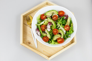 Fresh green salad with avocado, tomatoes, goat cheese in big white bowl on wooden tray. Vegan salad with green mix leaves  and vegetables. Top view on gray table. Healthy vegetarian food. Close up