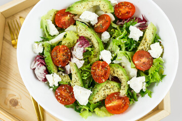 Fresh green salad with avocado, tomatoes, goat cheese in big white bowl on wooden tray. Vegan salad with green mix leaves  and vegetables. Top view on gray table. Healthy vegetarian food. Close up