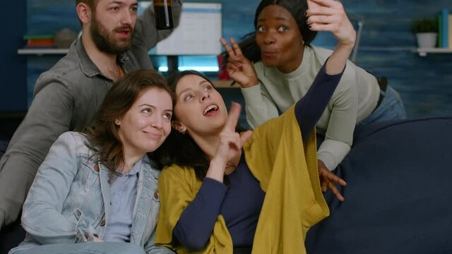 Multiracial friends taking sefie together using smartphone posting on social media. Group of mixed-race people sitting on couch late at night in living room enjoying free time