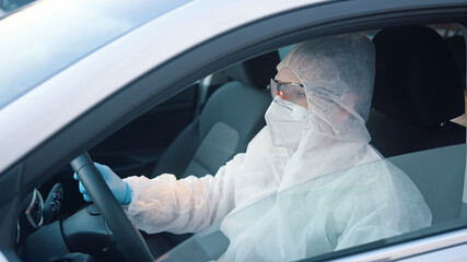 Unidentifiable person in protective overall suit driving car. Car side window view on man in infection risk area. High quality photo