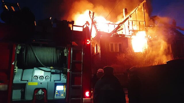 The roof of the house is on fire. The residential building burn, village. Firefighters put out a fire. Fire truck, headlights and panel, siren signal.