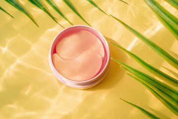 Pink jar with hydro gel eye patch on yellow background with palm leaves and water highlights....