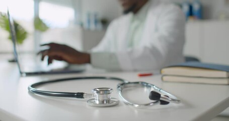 Focus on stethoscope lying on table and african doctor typing on laptop over blurred background