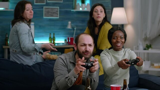 Woman with blak skin playing online videogames with man friends for gaming competition using controller. Group of mixed race winning game while sitting on sofa in living room late at night