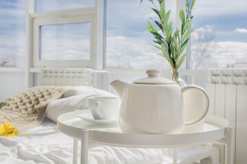 White teapot with tea, white cup stand on a white table in the morning in the bedroom