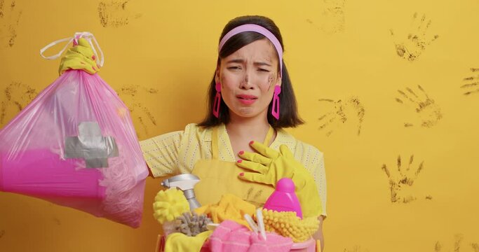 Displeased housewife looks with aversion at garbage bag cannot stand unpleasant stench poses near laundry basket wears domestic clothes stands against yellow background. Cleaning day concept