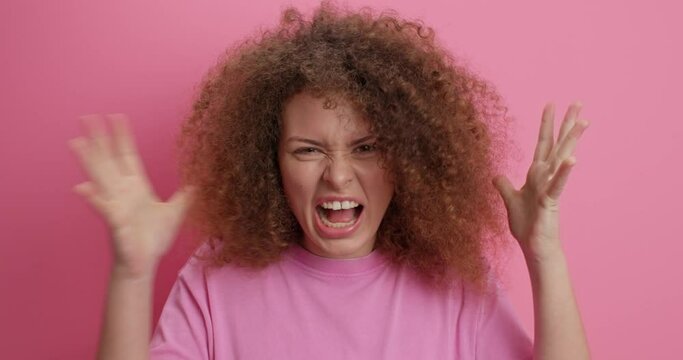 Emtional outraged woman with curly hair screams from anger shakes hands in dismay keeps mouth wide opened loses temper yess loudly expresses fury isolated over pink background. Dont bother me